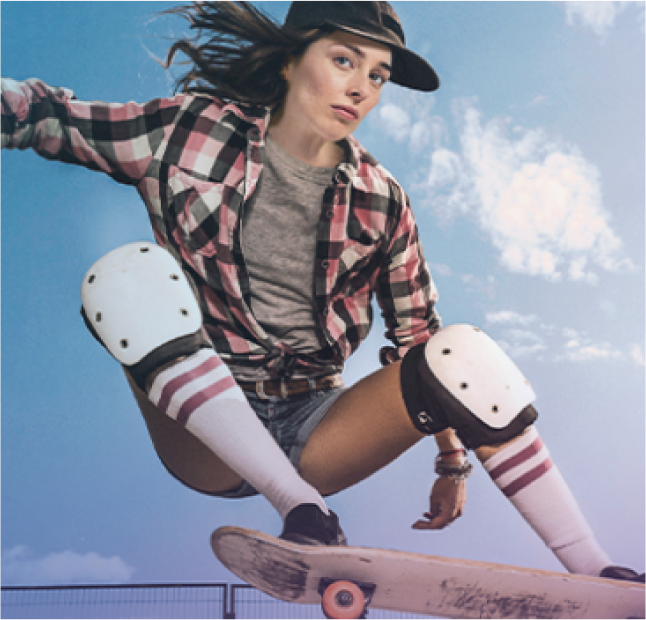 Frau, die auf einem Skateboard durch die Luft fliegt und Knieschoner, hohe gestreifte Socken, eine Kappe, ein langärmliges Flanellhemd und kurze Hosen trägt