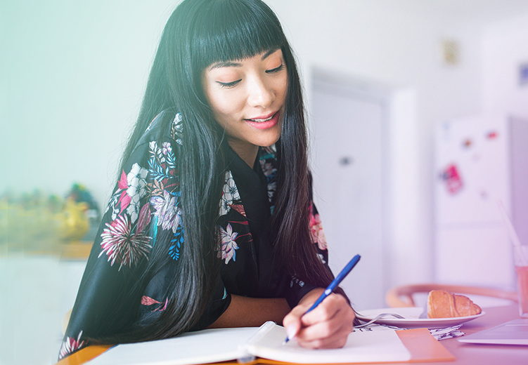 Femme aux longs cheveux foncés écrivant dans un cahier de notes tout en souriant.