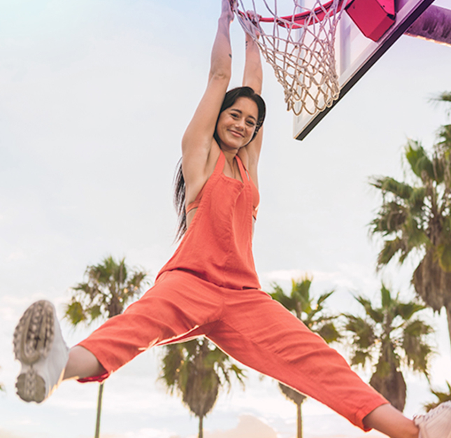 Femme souriante et vêtue d’une salopette orange suspendue à un anneau de basketball.