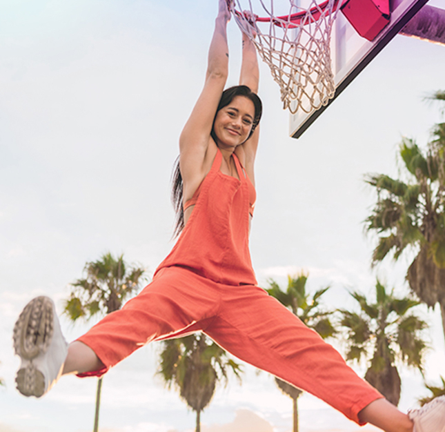 Woman in orange overalls hanging on basketball hoop rim smiling