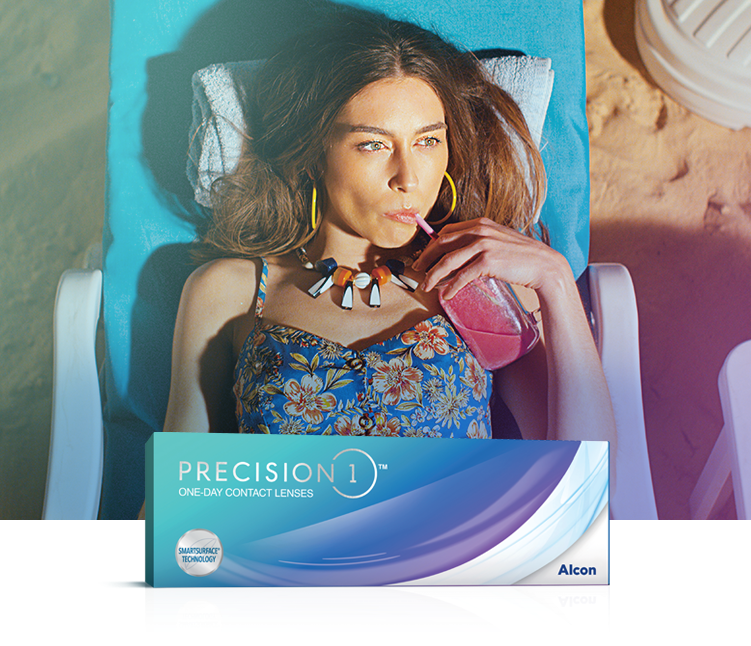 Woman with long brown hair in swimsuit sipping a pink colored drink while reclining on the beach—with Precision1 One Day Contact Lenses product box in foreground