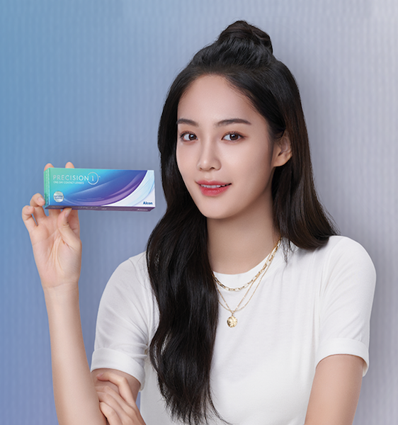 young woman with long hair wearing white shirt and gold chain necklaces holding a box of Precision1 One-Day Contact Lenses by Alcon