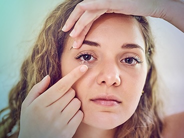 Close-up of a woman placing a contact lens into her eye, gently holding her upper eyelid while guiding the lens in with her finger.