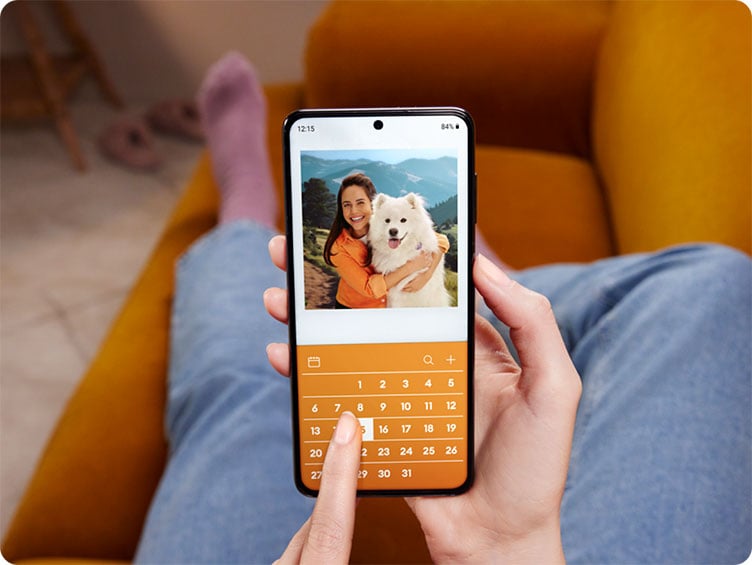 Person holding a smartphone while sitting on a couch, viewing a photo of a woman hugging a fluffy white dog, with a calendar interface below the image