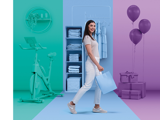 Mulher jovem sorridente a caminhar com confiança com um saco de compras azul-claro, passando por zonas coloridas com uma bicicleta de exercício, guarda-roupa e presentes com balões.