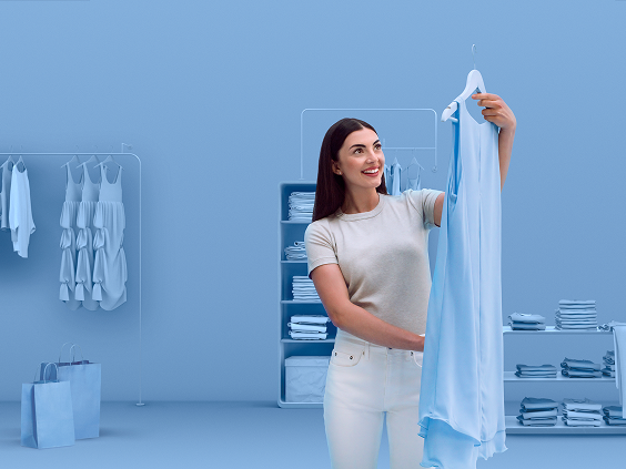 Mulher jovem a sorrir enquanto segura um vestido azul-claro num guarda-roupa elegante, vestida de forma casual e a aproveitar o momento, refletindo um estilo de vida moderno e confiante.