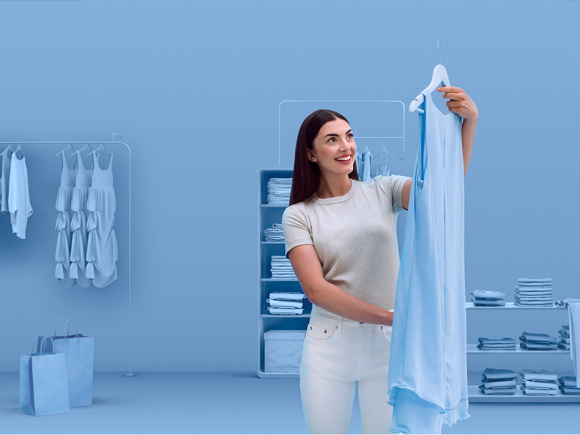 Smiling young woman holding up a light blue dress in a stylish wardrobe space, wearing a casual outfit and enjoying the moment, reflecting a confident, modern lifestyle.