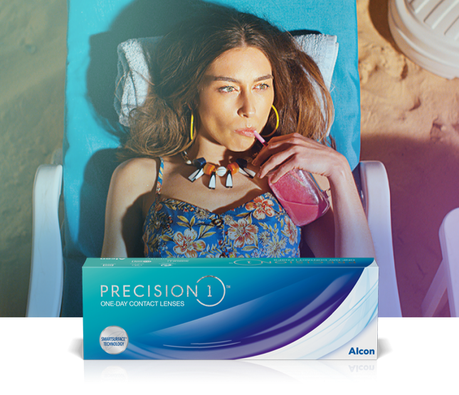 Woman with long brown hair in swimsuit sipping a pink colored drink while reclining on the beach—with Precision1 One Day Contact Lenses product box in foreground