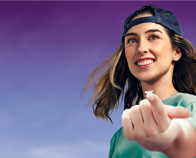 young woman with light brown hair balancing a contact lens on finger tip while smiling and wearing a cap backwards