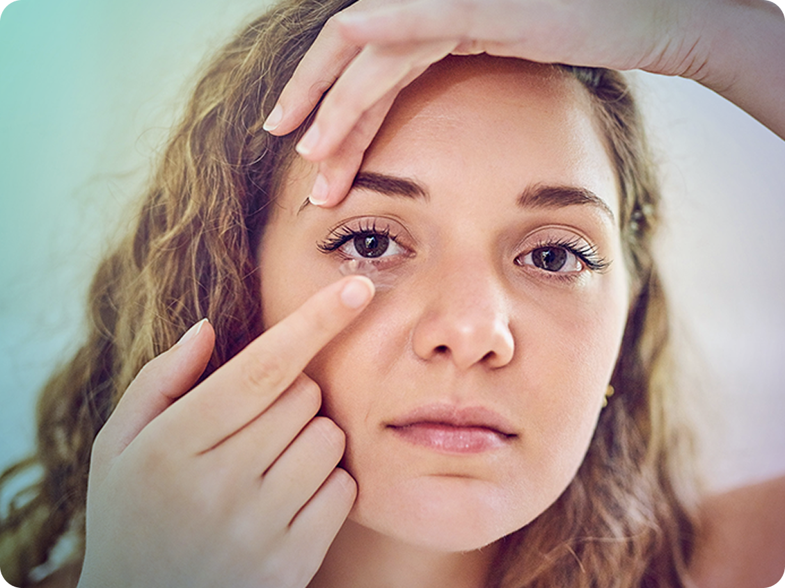 young woman with light brown hair balancing a contact lens on finger tip while smiling and wearing a cap backwards
